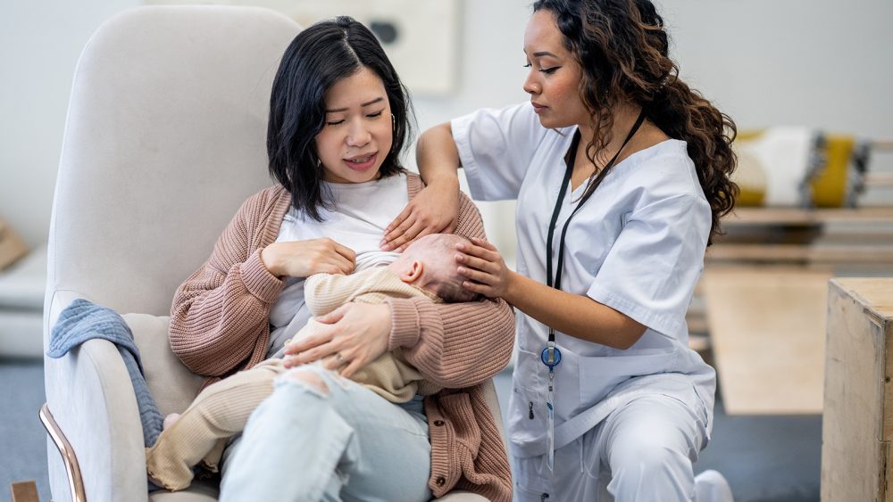 A new Mother attends a Lactation Consultant appointment as she looks for guidance with breastfeeding her daughter.  The practitioner is dressed professionally in scrubs as she calmly helps with positioning and latch.